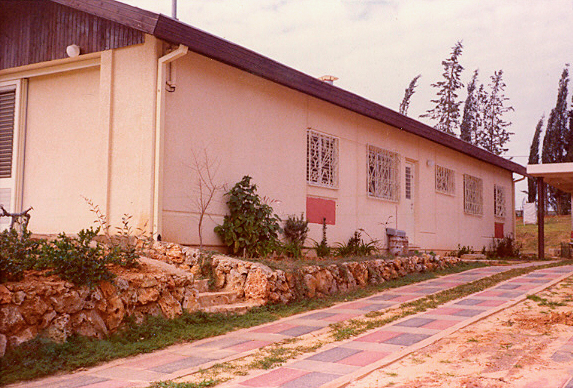 Side view of a single-story building with a stone pathway, surrounded by greenery and trees.